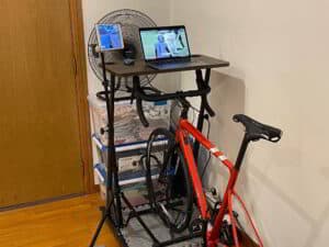 Red road bike on an indoor trainer beside a standing desk with a laptop